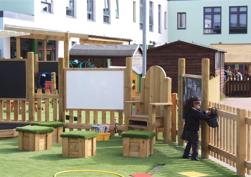 4 wooden boxes and a big wooden chair are placed around a whiteboard outside. The other side of the wooden chair is a chalkboard, with a child selecting chalk from a bucket. This area is fenced off by a small wooden fence.